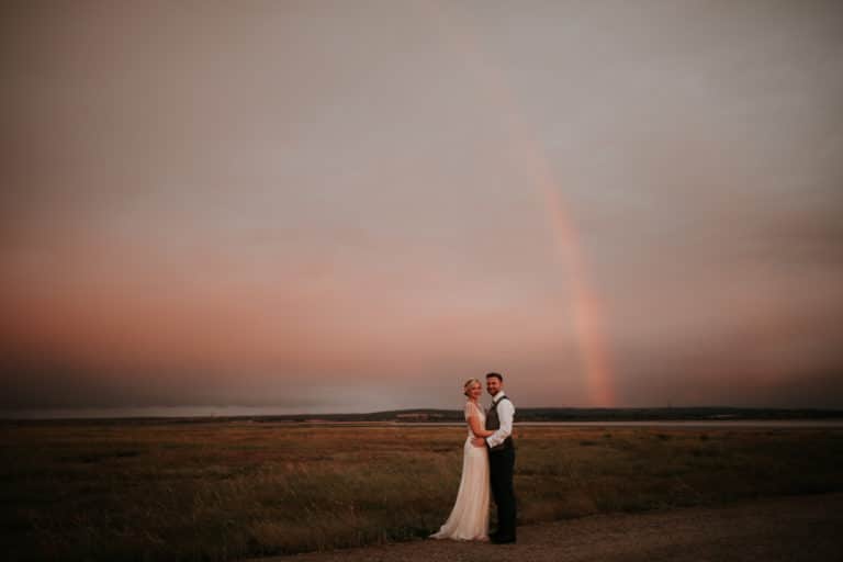 A wedding couple standing in the meadow at twilight, with a rainbow overhead