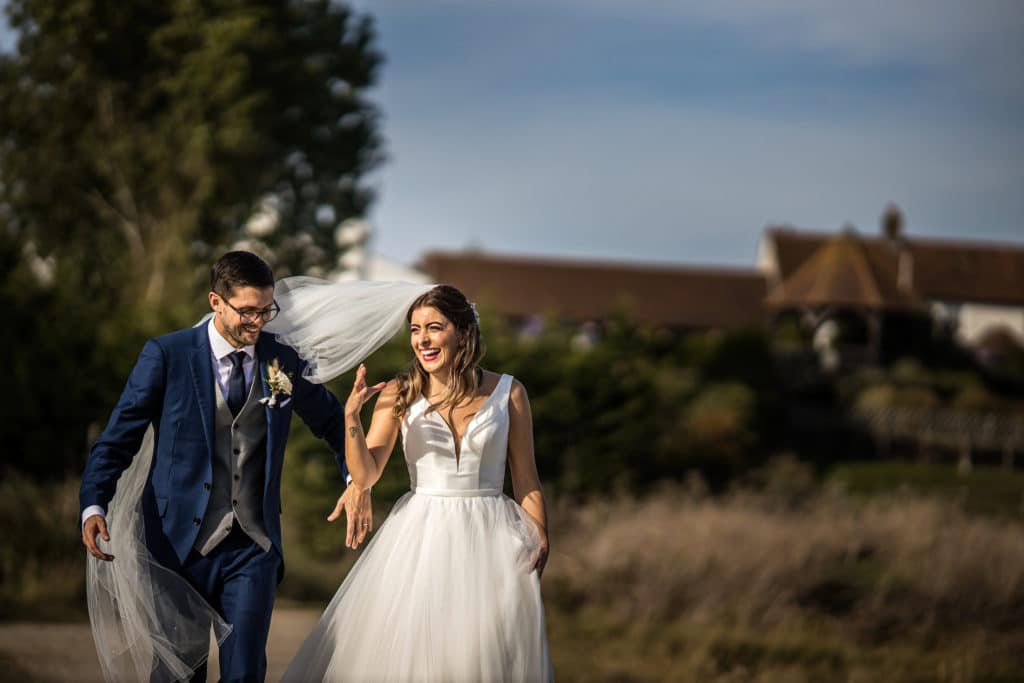 A wedding couple walking and laughing with The Ferry House wedding venue in Kent behind them