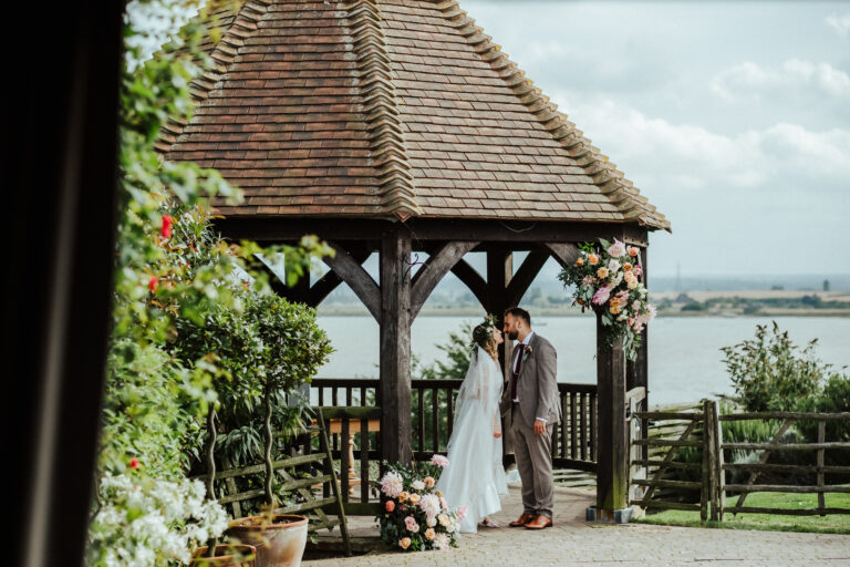 A wedding couple standing beside a gazebo overlooking the water, at The Ferry House barn wedding venue in Kent