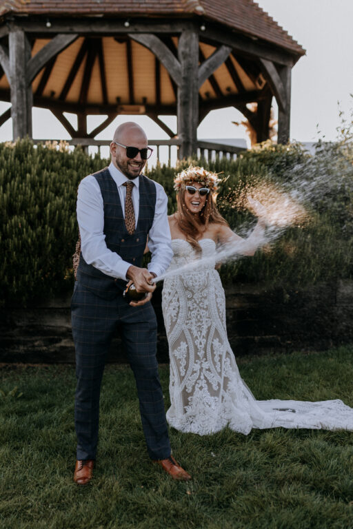 A bride and groom wearing sunglasses spraying a bottle of prosecco in the wedding gardens at The Ferry House.