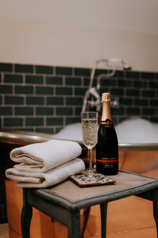 The copper bath in Room 6 at The Ferry House filled with bubbles with a glass of Chapel Down on a table.