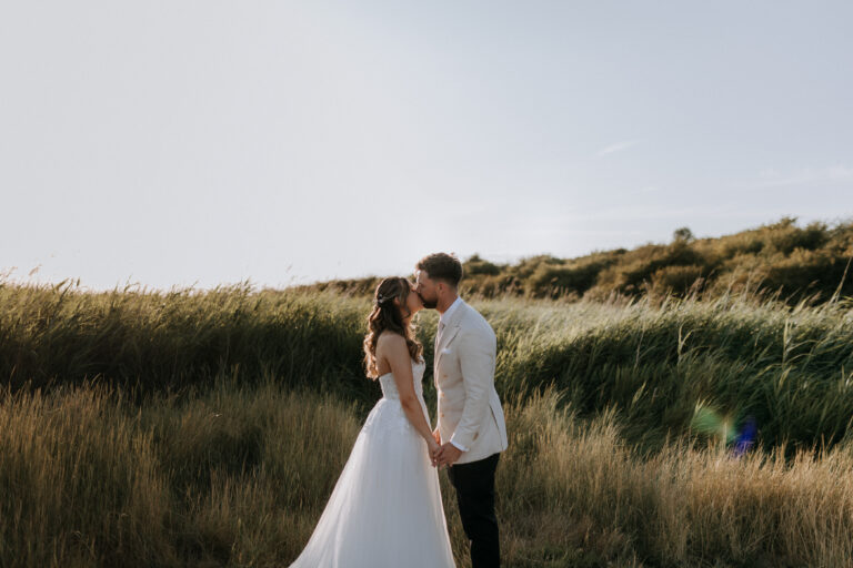 A bride and groom sharing an intimate moment on their wedding day at The Ferry House in Kent, with a backdrop of tall, wild grasses.