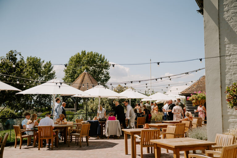 A wedding drinks reception from a sunny terrace with umbrellas and festoon lights.